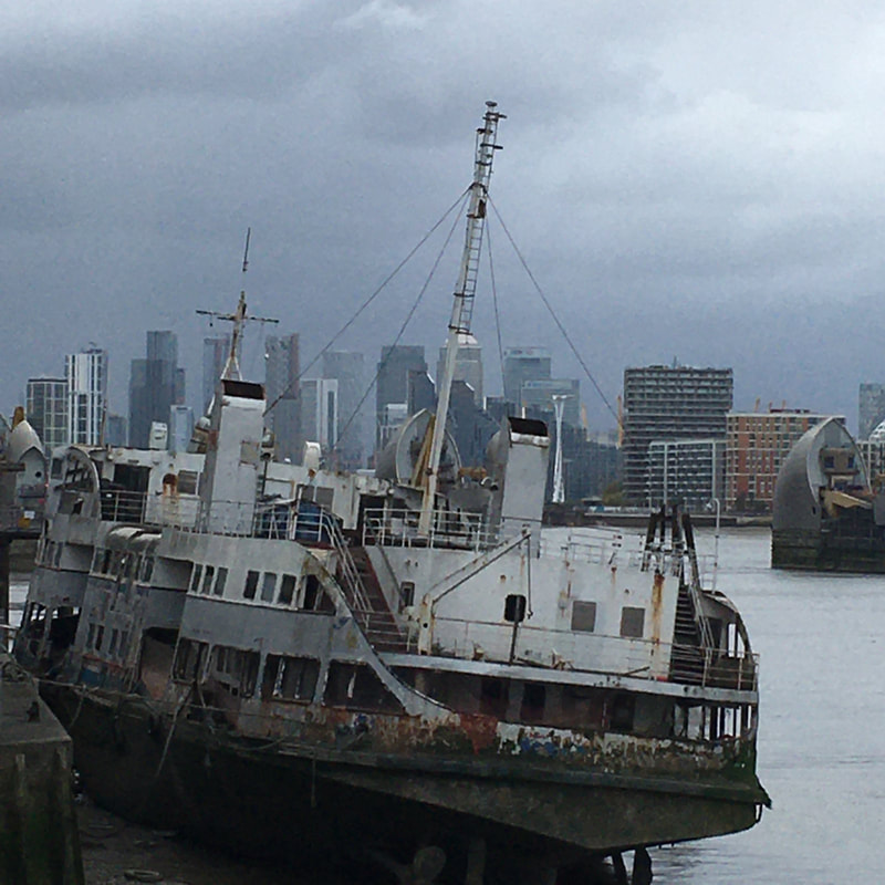 Picture: Rusting old de-commissioned large boat in foreground on the River Thames, with Thames Barrier in the mid, with cityscape of tall sky-scraper buildings of London in the background with moody and ominous grey clouds influencing the colour pallet of whole image of greys and light blues.