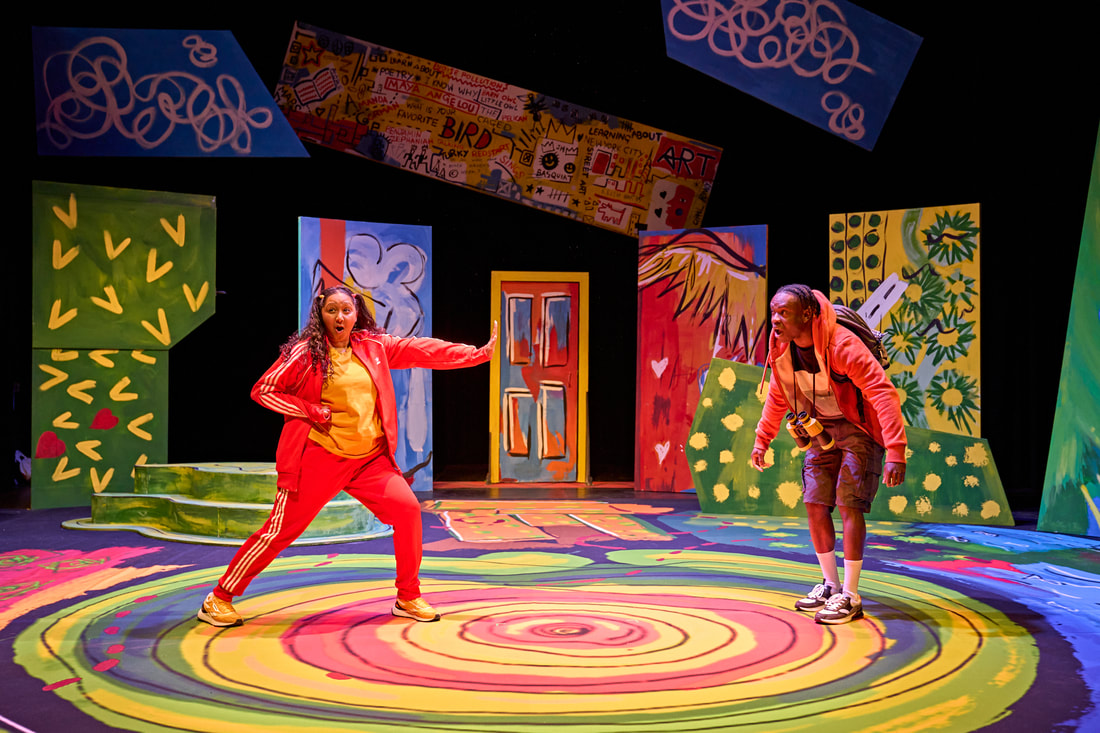 A wide angle image of the whole set of Marvin's Binoculars with two actors performing on it.  A young Asian woman is from a distance enacting a karate move using her hands in a punch towards the young Black African man who reacts by pretending he is getting punched in stomach.  She Wears a red Adidas tracksuit with yellow trainers and a yellow t - shirt.  He wears black and white trainers, grey shorts, a black, grey and white t-shirt, an orange unzipped hoody, yellow and black binoculars around his neck and a rucksack.  The set is colourful in greens reds, yellows and blues.  On the floor a large series of concentric circles alike the inner of a tree trunk; a door central at the back. Upright painted set pieces, including two that looks like trees; one a bush.  Two abstract flats one in blue and one in red flank the door, both have yellow wings painted on. Suspended above towards the back two pieces that give impression of sky and white clouds and a long yellow display board with graffiti style writing with names of authors such as Maya Angelou and artists such as Nina Chanel Abney.