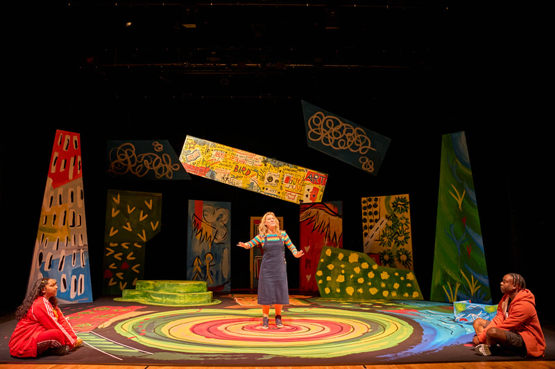 A white woman centre stage wearing a blue dress with a colourful stripey top, orange tights and blue high top boots, hands outstretched as if to quieten those listening.  The set in the background includes colourful set 'flat's, to the left a piece that suggests a tree, a blue sky with clouds to the left and right of the image; centrally, a display board with strong yellow colouring and writing in a grafitti style in black and red suspended in the air with a black background; to the left and behind the woman, a flat which is blue with a wing in yellow and to the doors right a flat which is red with a yellow wing.  On the ground a green mound with raised levels and to the right another flat to resemble a bush.  On the floor colourful concentric circles from outer to inner in greens, yellows, and reds alike the rings of a tree trunk when cut in half.  Other areas to indicate water in blue and earthy red to either side.  A playful scene with a childlike quality to the set.