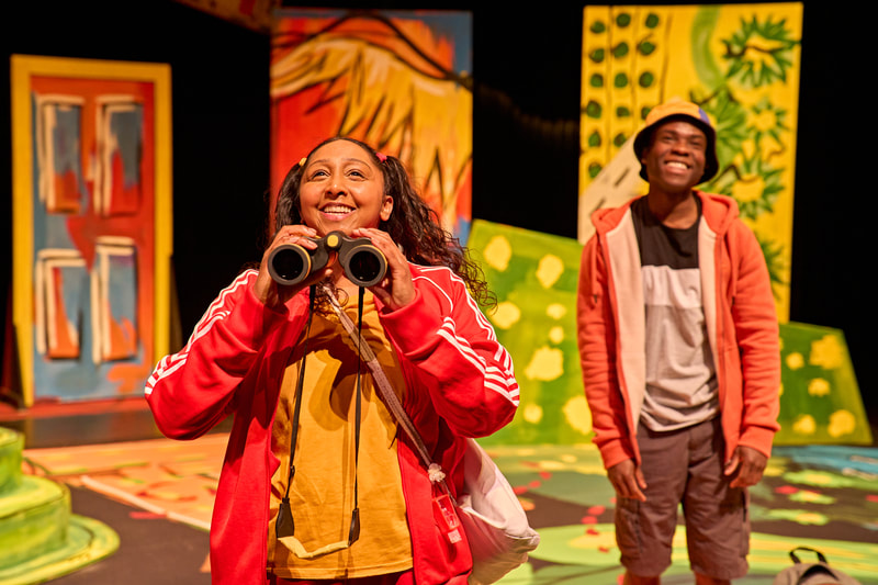 A young Asian woman with black curly hair wears a red Adidas tracksuit and yellow t-shirt.  She holds binoculars that are yellow and black in her hands as if she is about to use them.  In the background, a young black African man looks on with a smile. 
 He wears a colourful sun hat, orange hoody top that is unzipped, a black, white and grey t-shirt and grey shorts.  There are colourful set pieces of greens, yellows, red and yellow in the background.