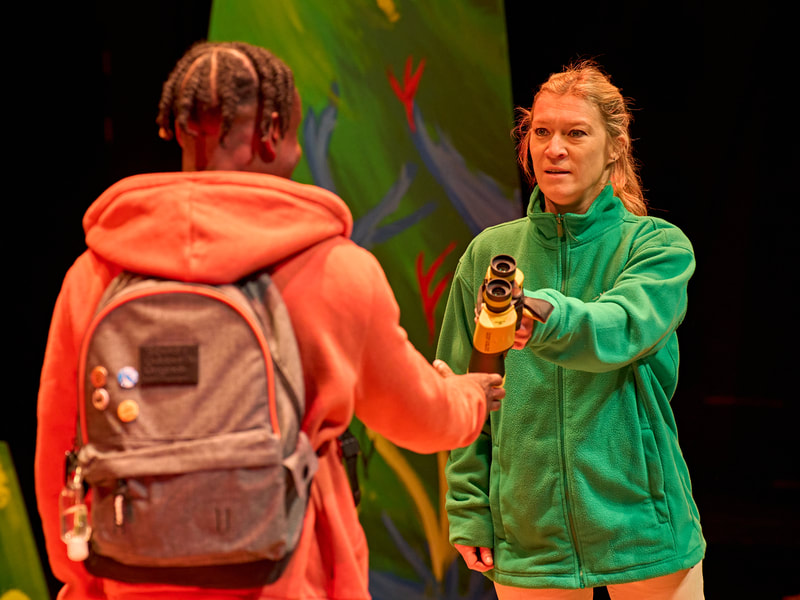 A white woman, wearing a green fleece holds out binoculars towards a black African young man whose back is to us.  He wears an orange hoody and grey rucksack with small badges on it.  He reaches towards the binoculars.