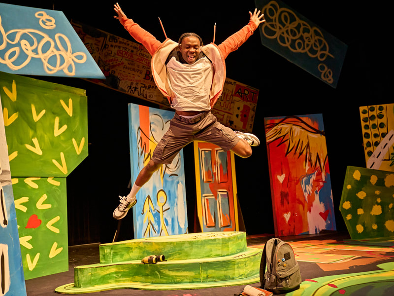 An African young man aged in his 20's leaping in a star jump.  He is wearing trainers; white socks, grey cargo shorts, a white black and grey t shirt with a orange zip up hoody. The set in the background includes colourful set 'flat's, to the left a piece that suggests a tree, a blue sky with clouds to the left and right of the image; a display board with strong yellow and writing in a grafitti style in black and red suspended in the air with a black background; a blue, red and yellow door and to its left a flat which is blue with a wing in yellow and to the doors right a flat which is red with a yellow wing.  On the ground a green mound with binoculars set on a step. A grey rucksack set on the floor.  