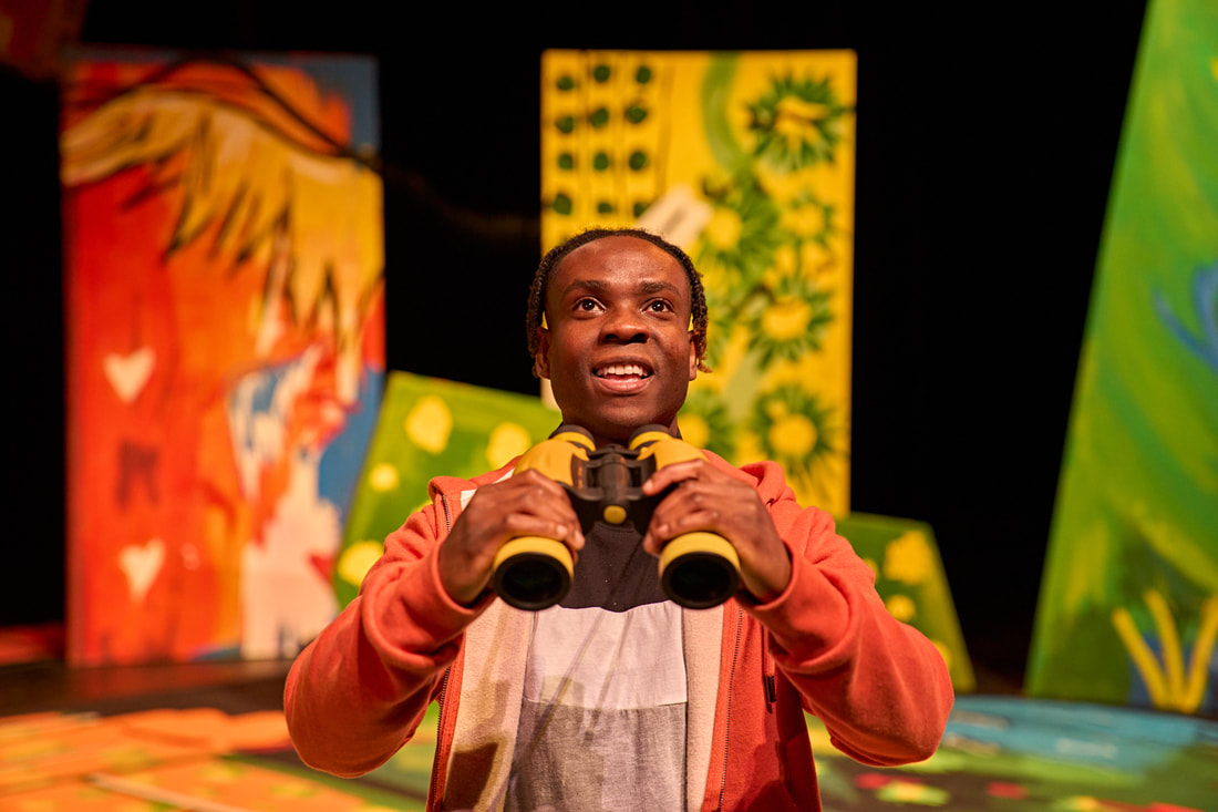 A young black African man looks on with a smile. He is holding in both hands as if to look through them - yellow and black binoculars. He wears an orange hoody top that is unzipped with a black, white and grey t-shirt showing underneath.  There are colourful set pieces of greens, yellows, red and yellow in the background.