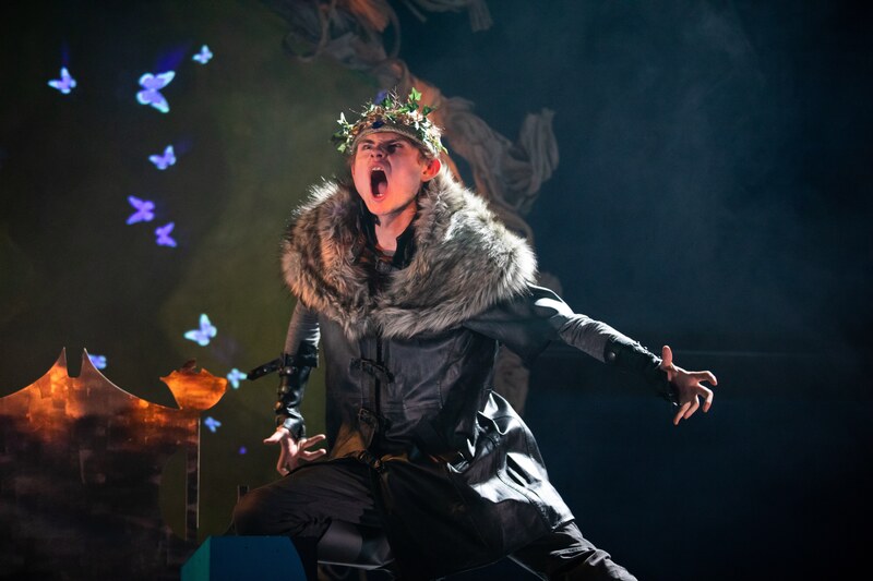 A young white man in his early twenties, arms outstretched to his sides with hands in in claw shape, he makes a roaring gesture with his face.  Wearing a crown of leaves, a faux black leather coat with greyish fur around the neck and shoulders, Wearing black trousers and legs in a aggressive ready to pounce stance.  He stands on a stage with a throne to his stage right and a projection of blue butterflies in the background.