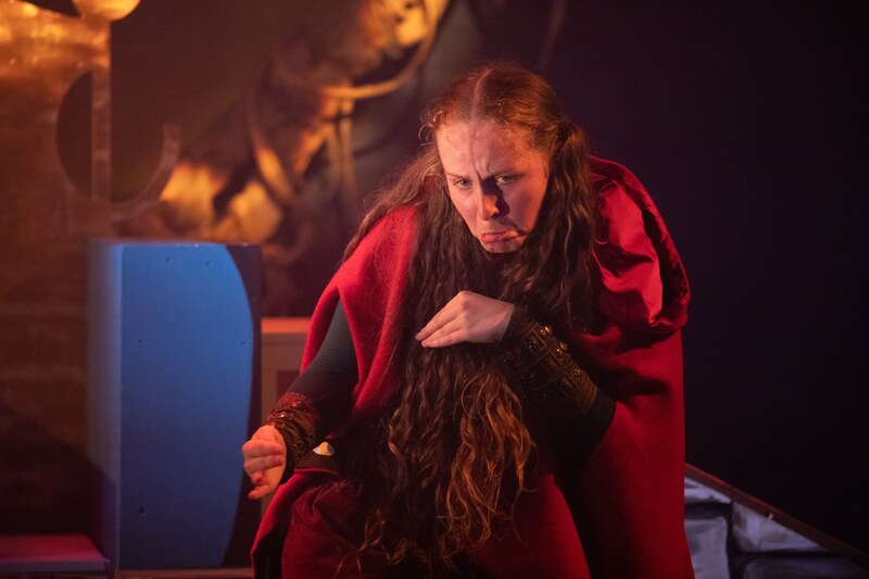 In the foreground, a young white woman with long hair with a a centre parting falling in front of her chest, wearing a red cloak retreating in an animal like stance with her left hand held cupped close to her chest like a paw.  In the background, blues from the set and golden blurred colours.