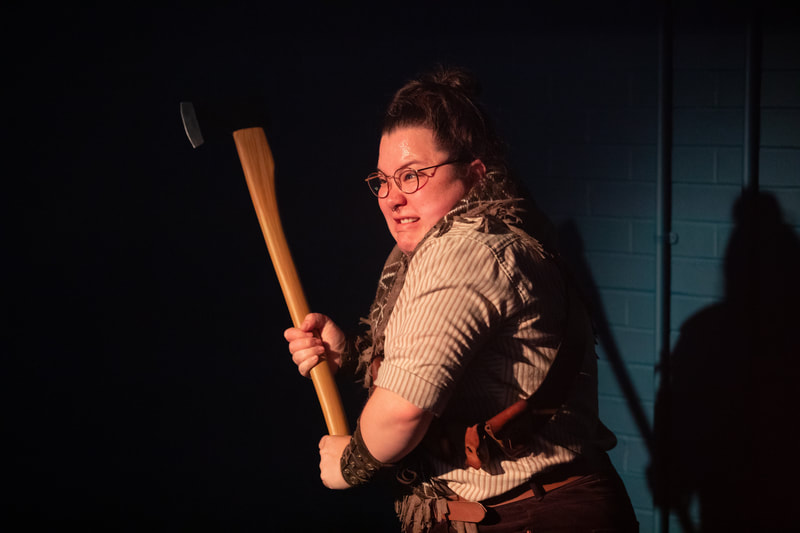 The person is holding an axe and is in an attacking position with an aggressive scowl upon their face.  They wear round glasses, a brown shawl and a light brown stripey shirt with rolled up sleeves. Their hair is pulled off their face. They are of mixed white and South Asian heritage. 